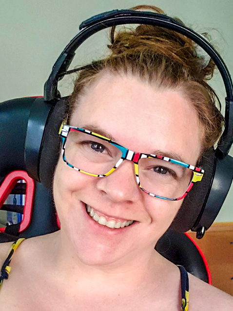 A smiling woman with brown hair, wearing black headphones and multicolored glasses.