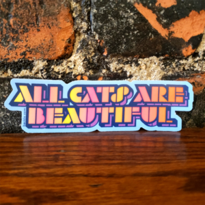 "All Cats Are Beautiful" sticker in an all caps stencil font. The letters have a dark blue outline with a gradient inside that has yellow, orange, and pink tones. Text has a purple shadow. Background is light blue.